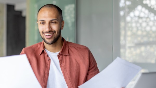 Smiling man in a rust-colored shirt holding and reviewing papers in a bright, modern office