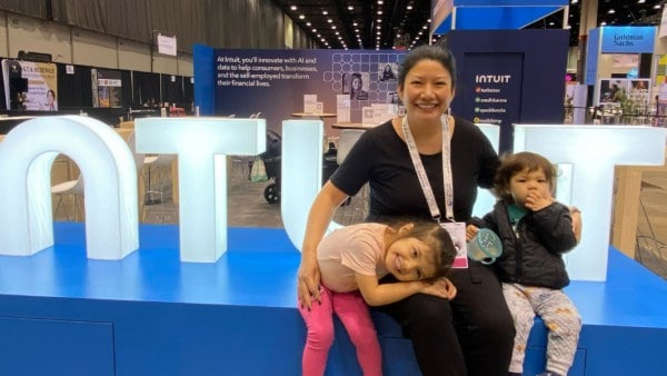 Woman smiling with two young children sitting in front of large ‘INTUIT’ letters at an Intuit booth in a busy convention expo hall