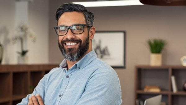 Smiling man with glasses and a beard standing with arms crossed in a modern office