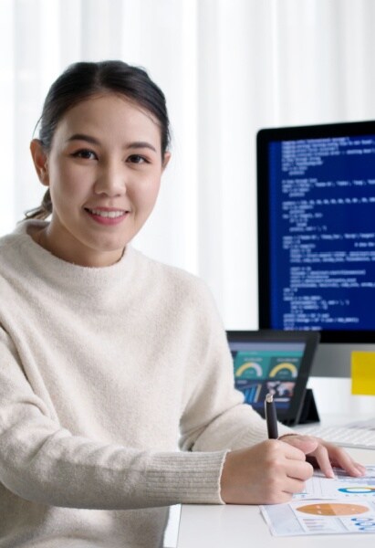 Smiling woman sitting at a desk with a computer displaying code and data charts in a bright office