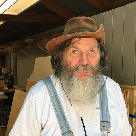 A person in a brown hat with a beard wearing white t-shirt and overall 