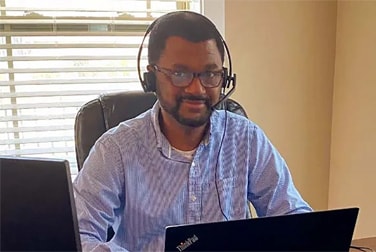 A person wearing glasses and a blue shirt is sitting at a desk with two laptops.